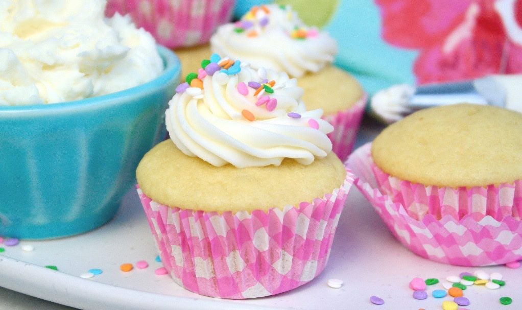 Vanilla cupcakes with white frosting and colorful sprinkles in pink wrappers, next to a blue bowl of whipped cream.