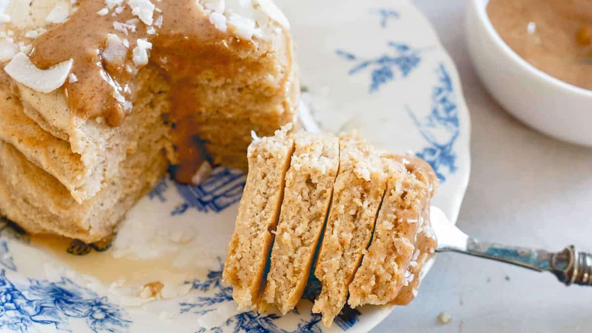 A close-up of a stack of pancakes with syrup and shredded coconut on a blue and white plate, with a fork holding a cut piece.