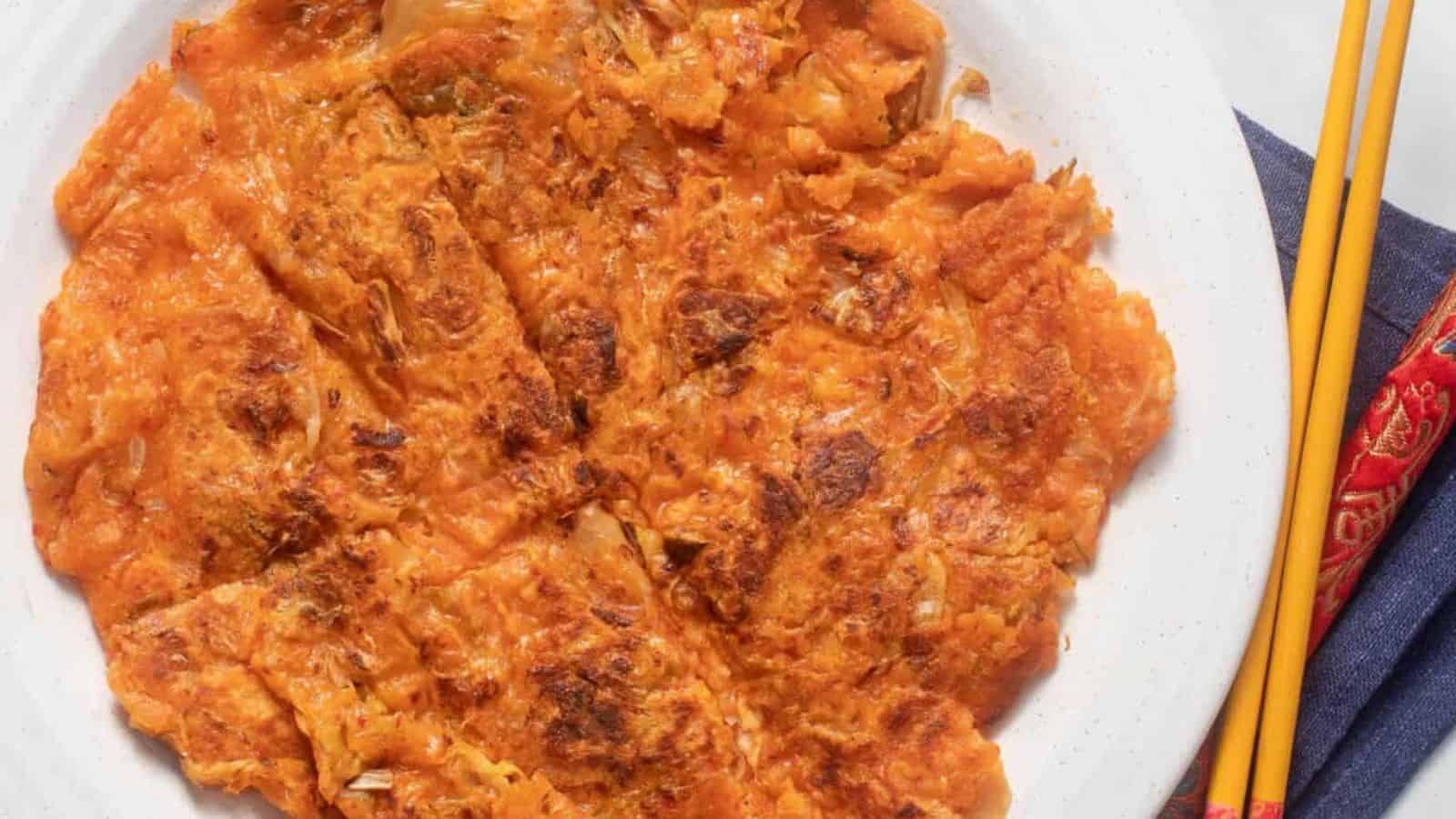 A plate of kimchi pancake, golden and crispy, sliced into pieces, with a pair of chopsticks placed beside the plate.