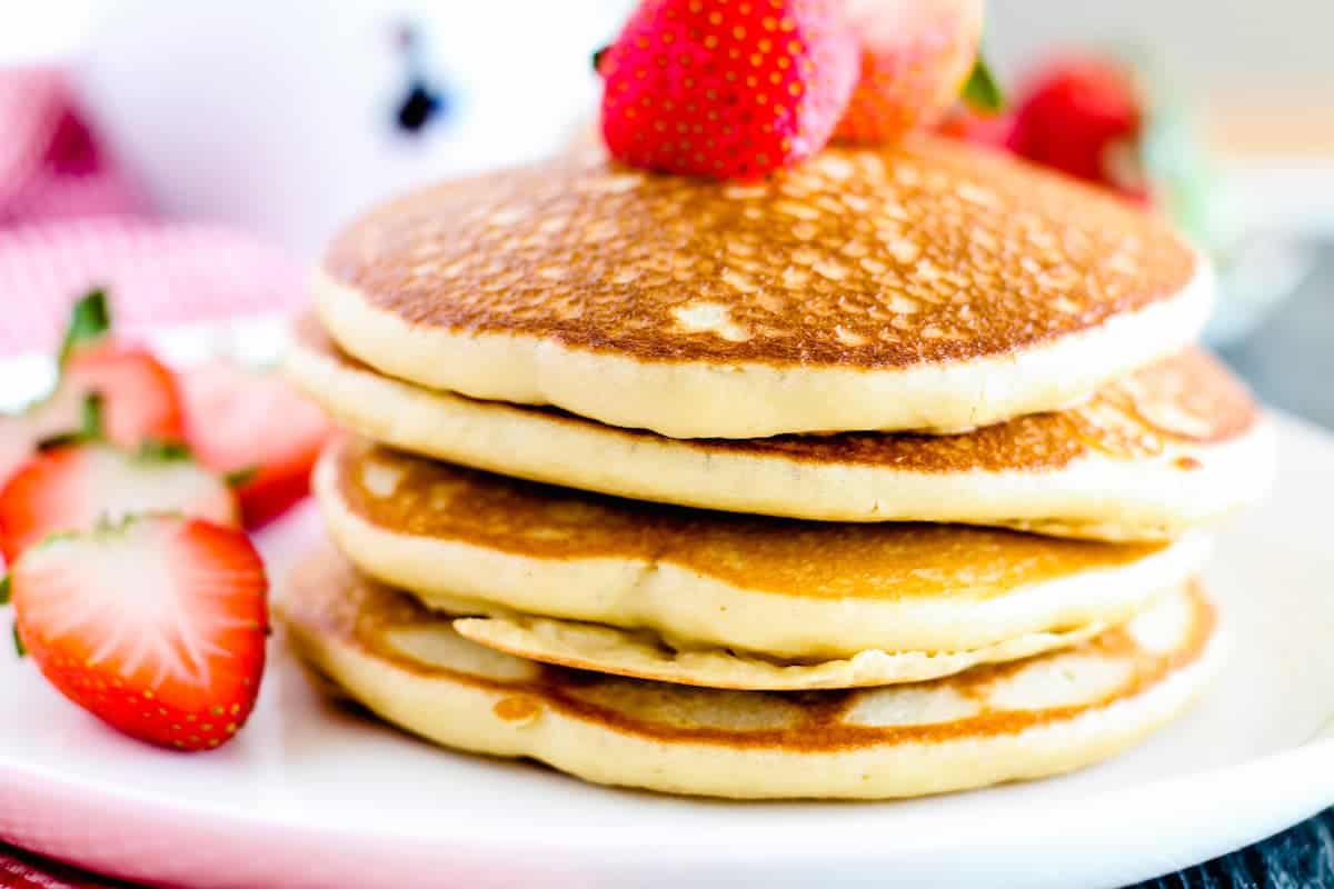 A stack of three pancakes topped with strawberries is served on a white plate, with sliced strawberries on the side.