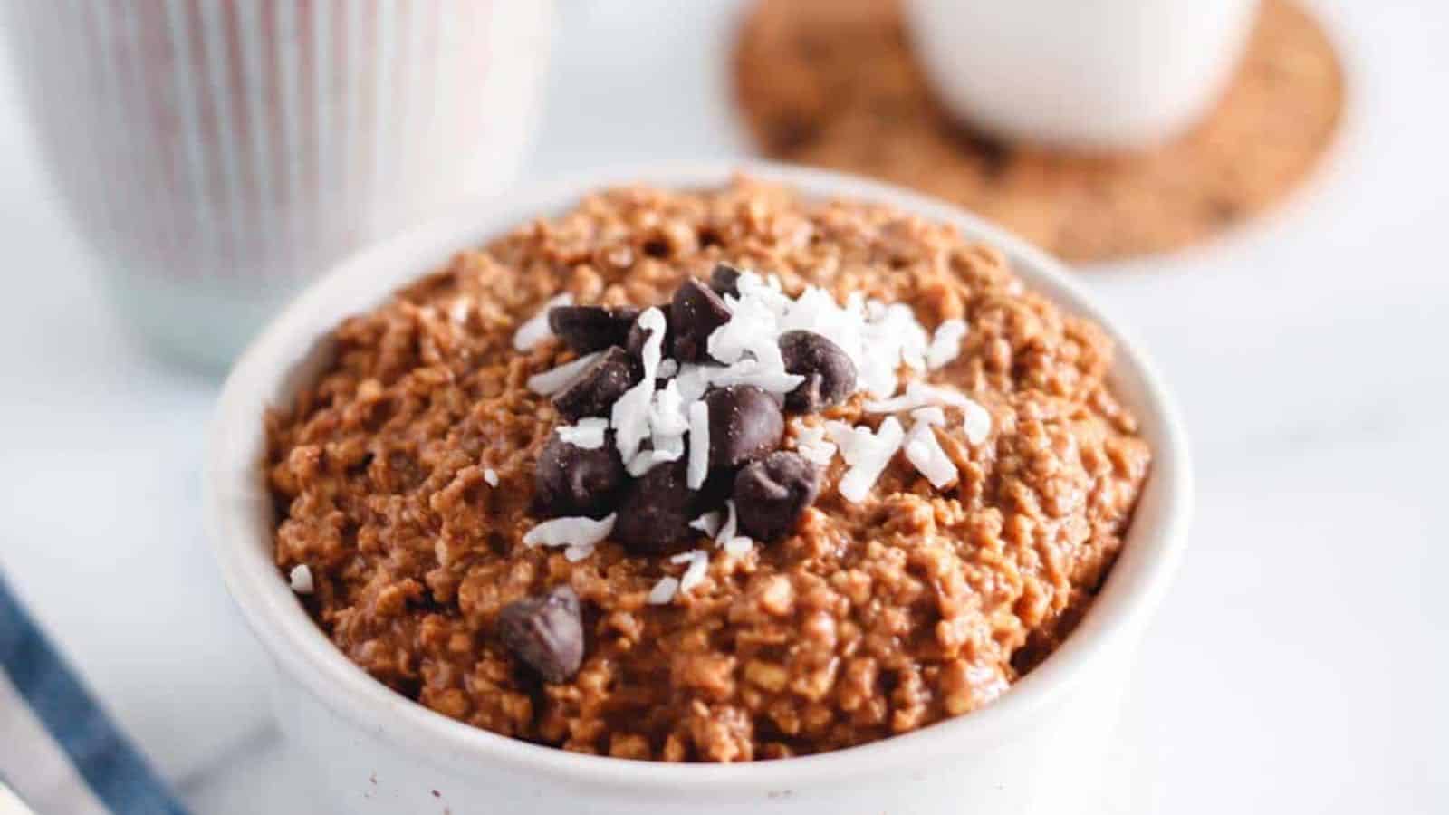 A bowl of chocolate oatmeal topped with shredded coconut and chocolate chips, placed on a white surface.