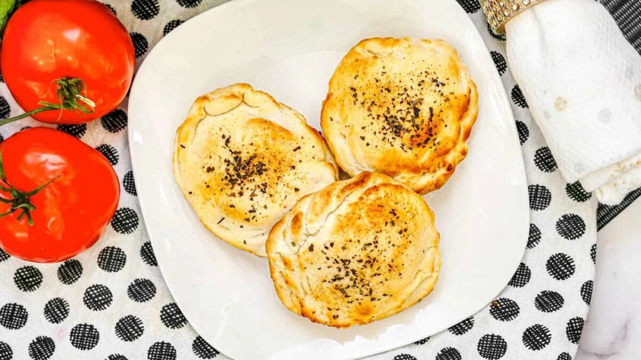 Three small, round savory pies topped with herbs are arranged on a white plate, next to two tomatoes, on a black and white polka dot tablecloth.
