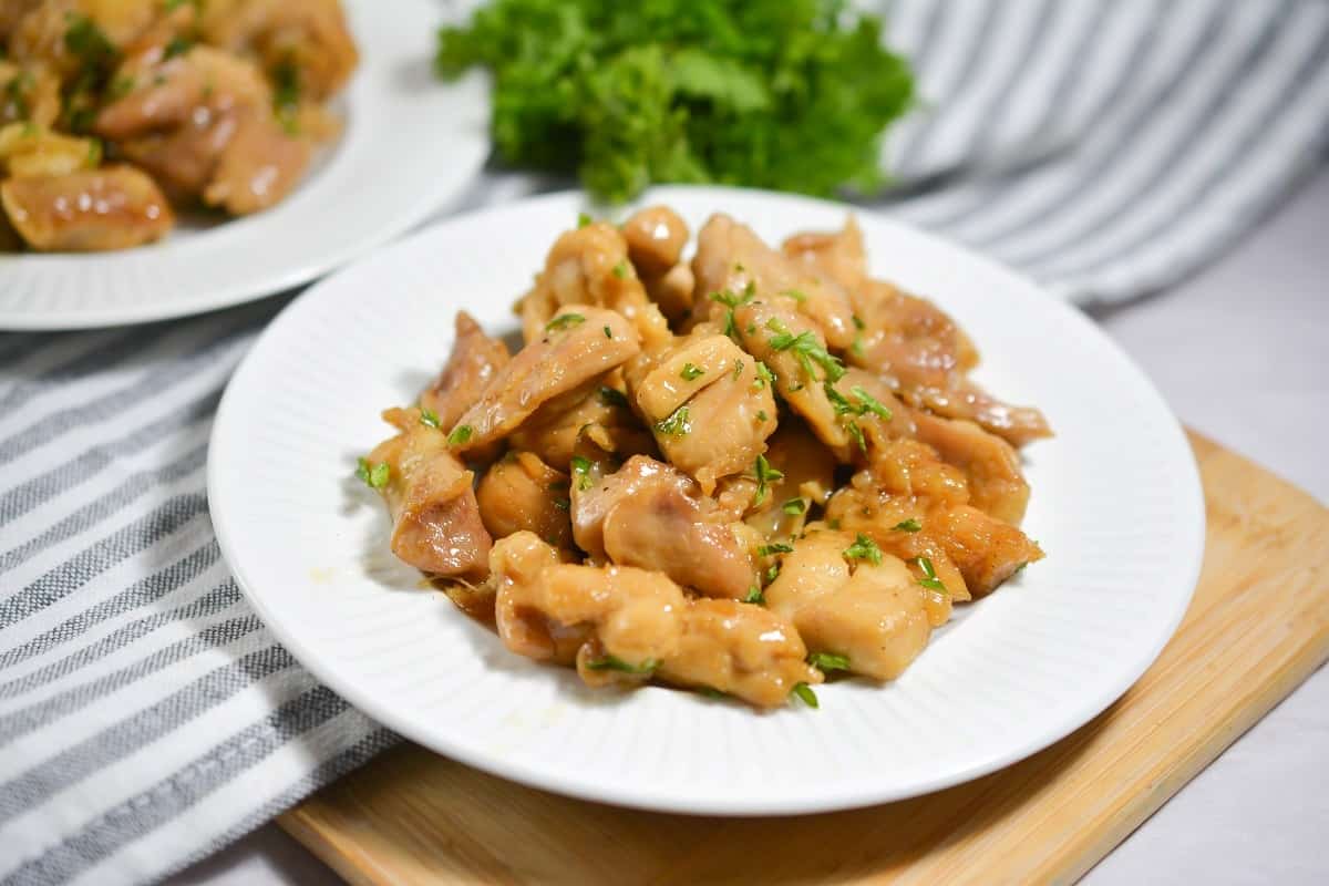A white plate of cooked chicken pieces garnished with chopped herbs, placed on a striped cloth with leafy greens in the background.