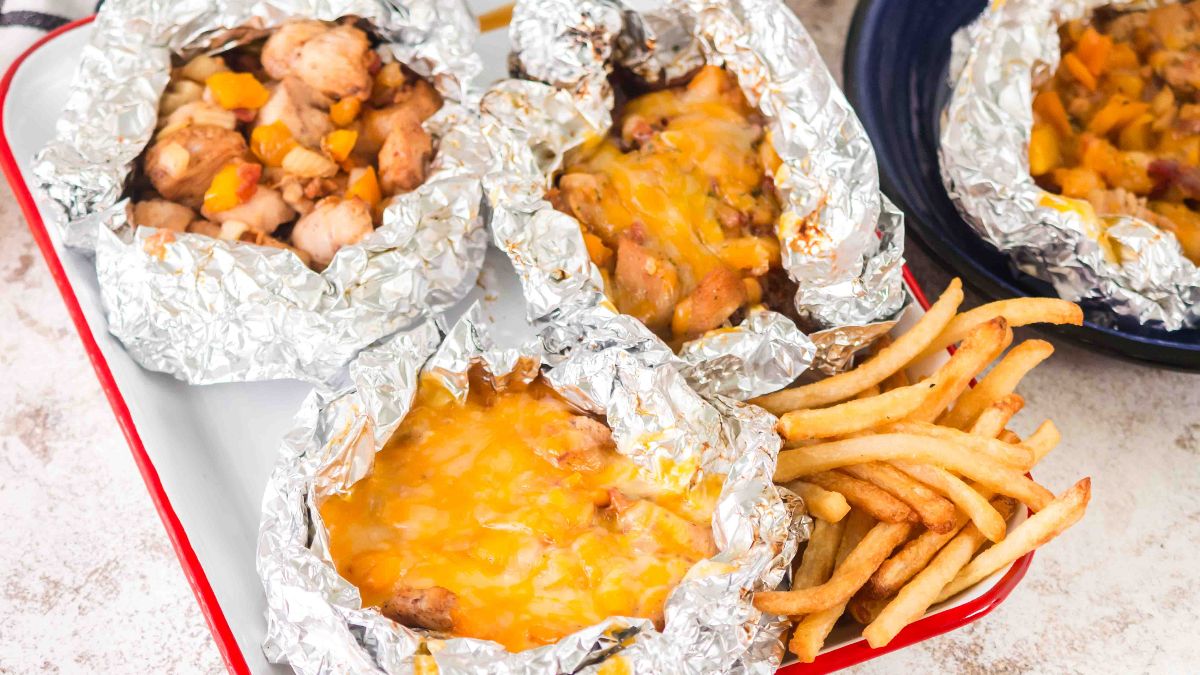 Four foil packets filled with cooked chicken and melted cheese on a tray, served with a side of French fries.