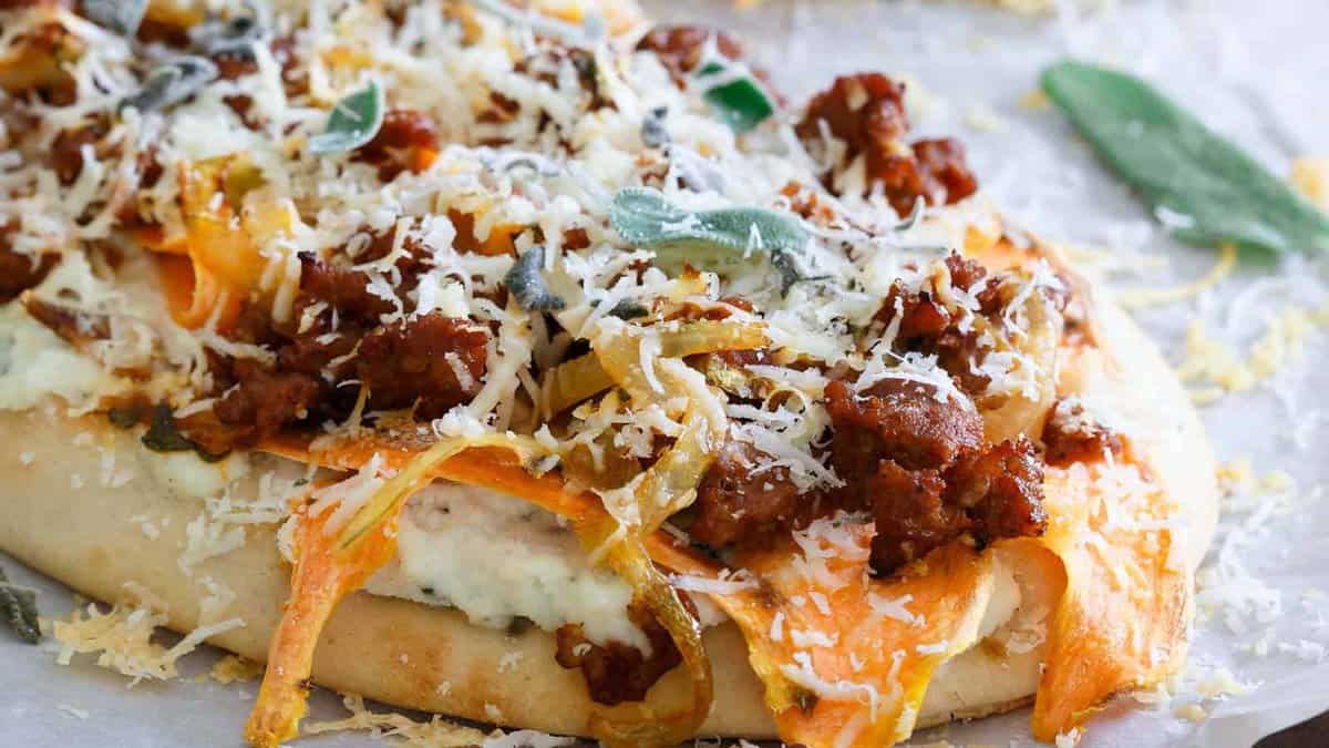 Flatbread topped with ground sausage, sliced butternut squash, caramelized onions, fresh sage leaves, and grated cheese on a parchment-lined surface.
