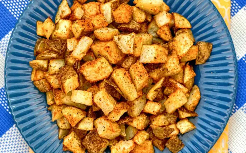 A blue plate filled with seasoned, roasted diced potatoes, placed on a checkered tablecloth.