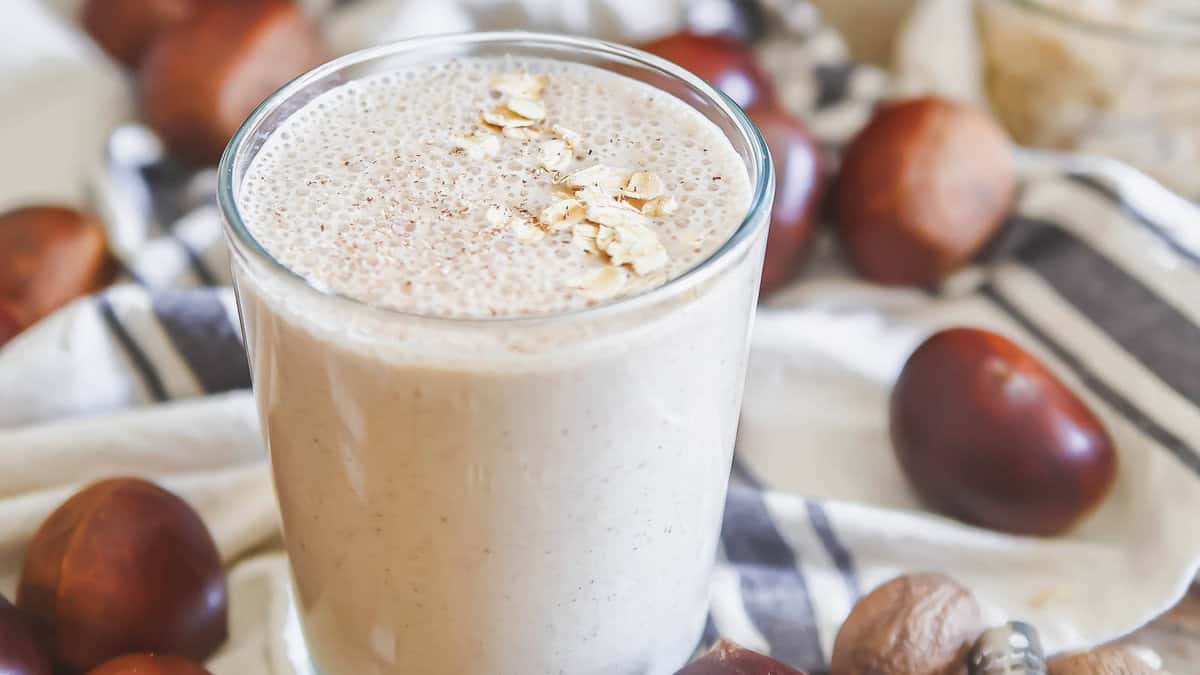 A glass of creamy smoothie topped with rolled oats, surrounded by whole chestnuts on a striped cloth.