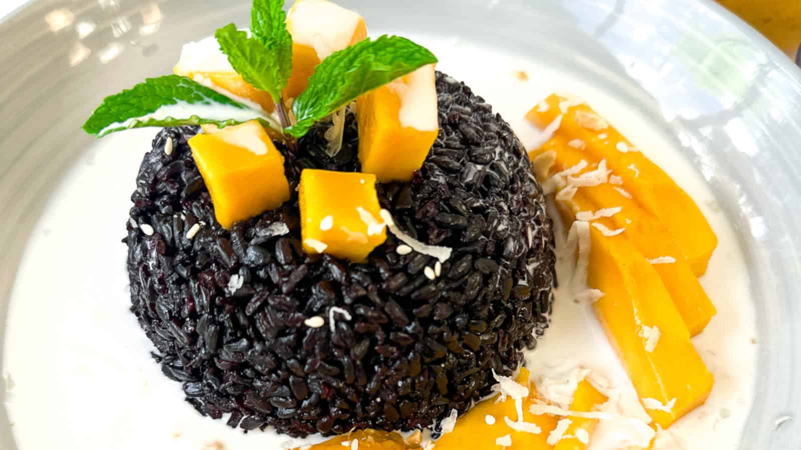 A serving of black sticky rice topped with mango cubes, shredded coconut, and mint leaves, plated with coconut milk and mango slices on the side.