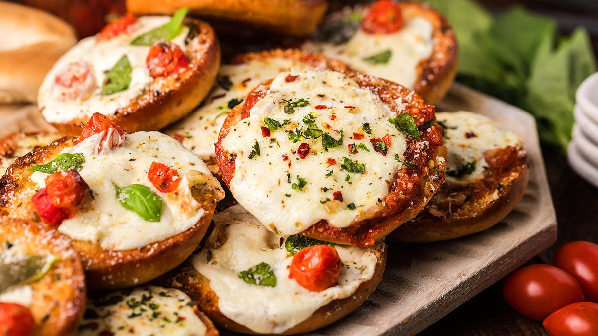 A close-up of several mini bagels topped with melted cheese, tomato pieces, and basil, arranged on a wooden surface.
