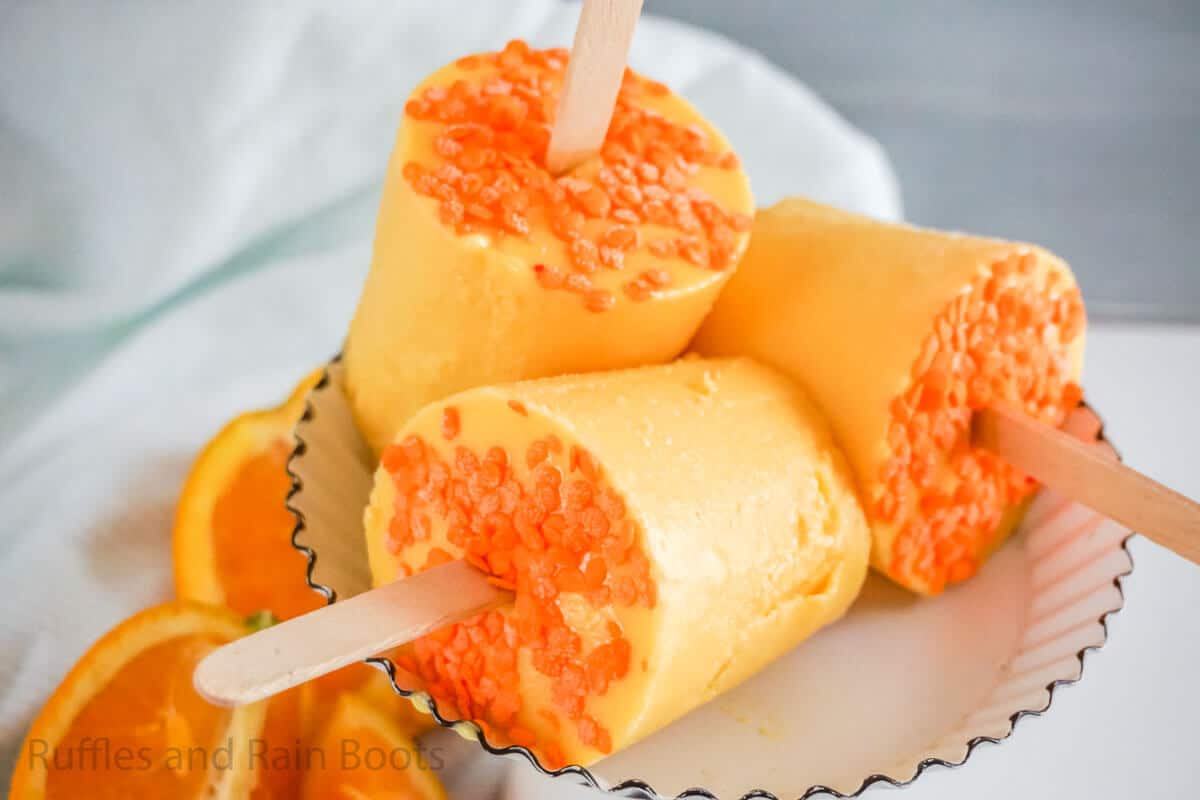Three orange popsicles with orange sprinkles on the sides are stacked on a white plate, with orange slices in the background.