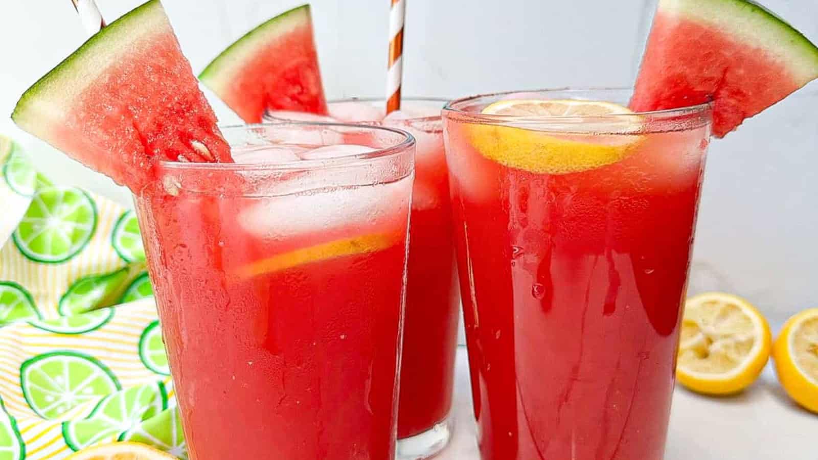 Three glasses of watermelon juice with ice, garnished with lemon slices and watermelon wedges; striped straws are inserted in each glass.