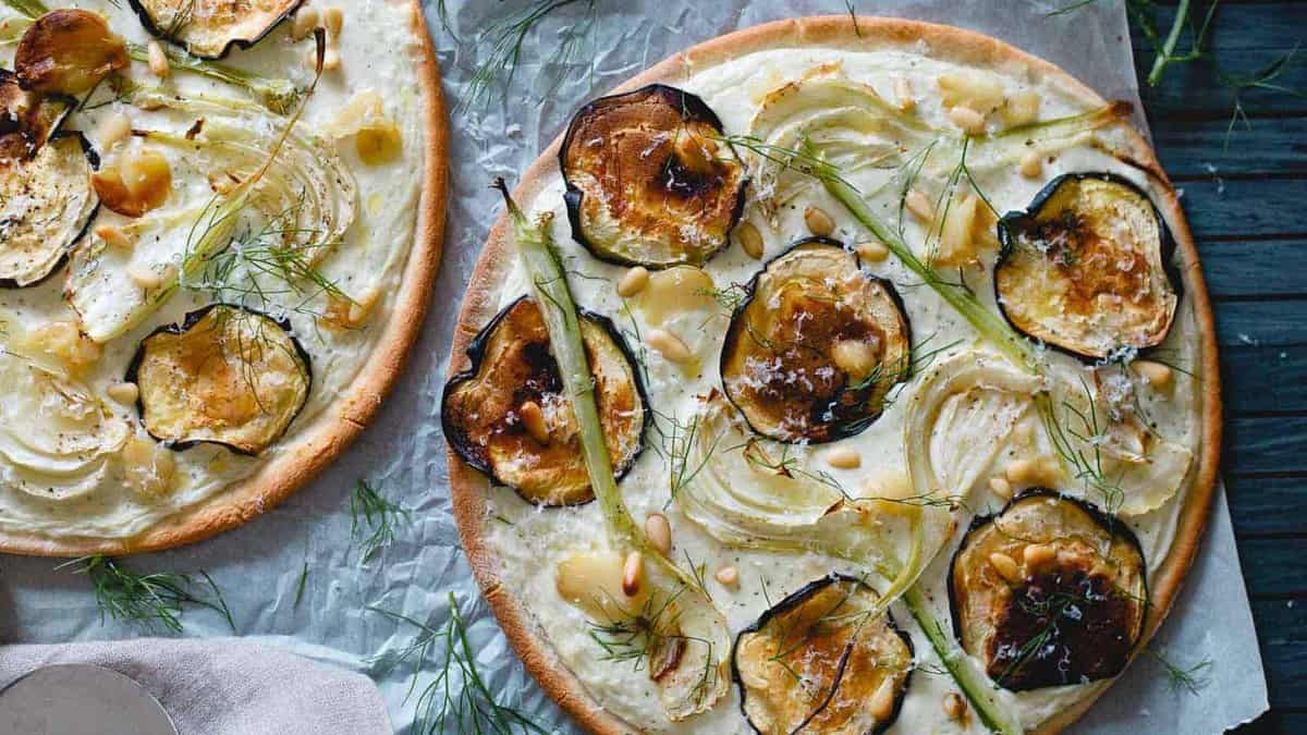 Two thin-crust pizzas topped with sliced zucchini, fennel, pine nuts, and cheese, garnished with fresh fennel fronds on parchment paper.