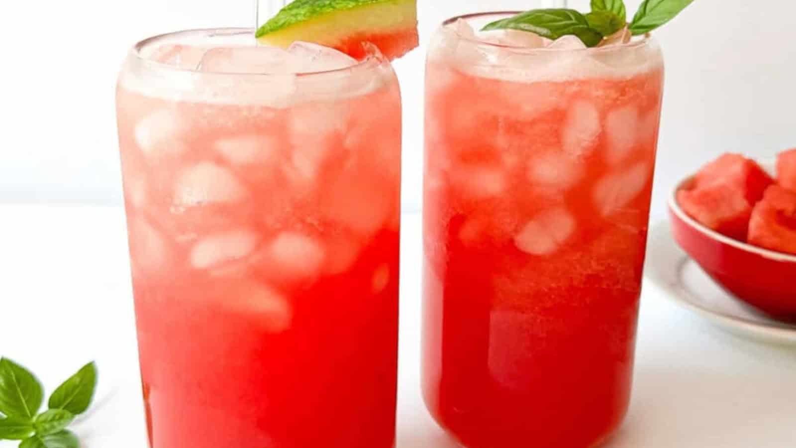 Two tall glasses filled with ice and red watermelon drink, garnished with watermelon slices and basil leaves. A small bowl of watermelon pieces sits in the background.