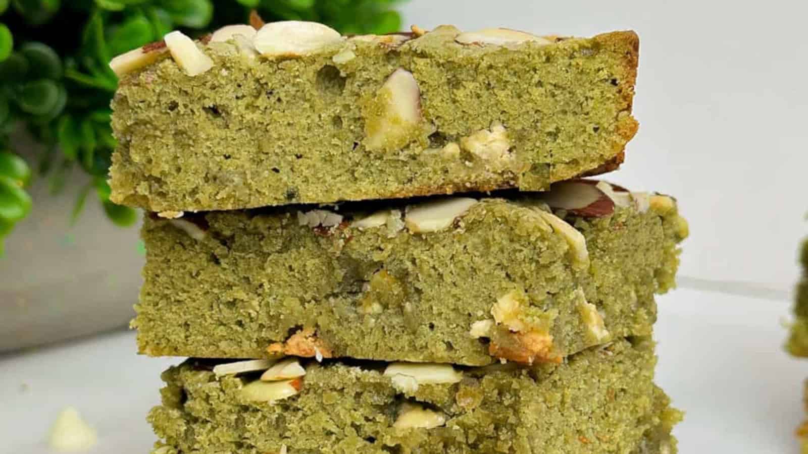 Three pieces of green cake with nut slices on top are stacked on a white surface, with a plant in the background.