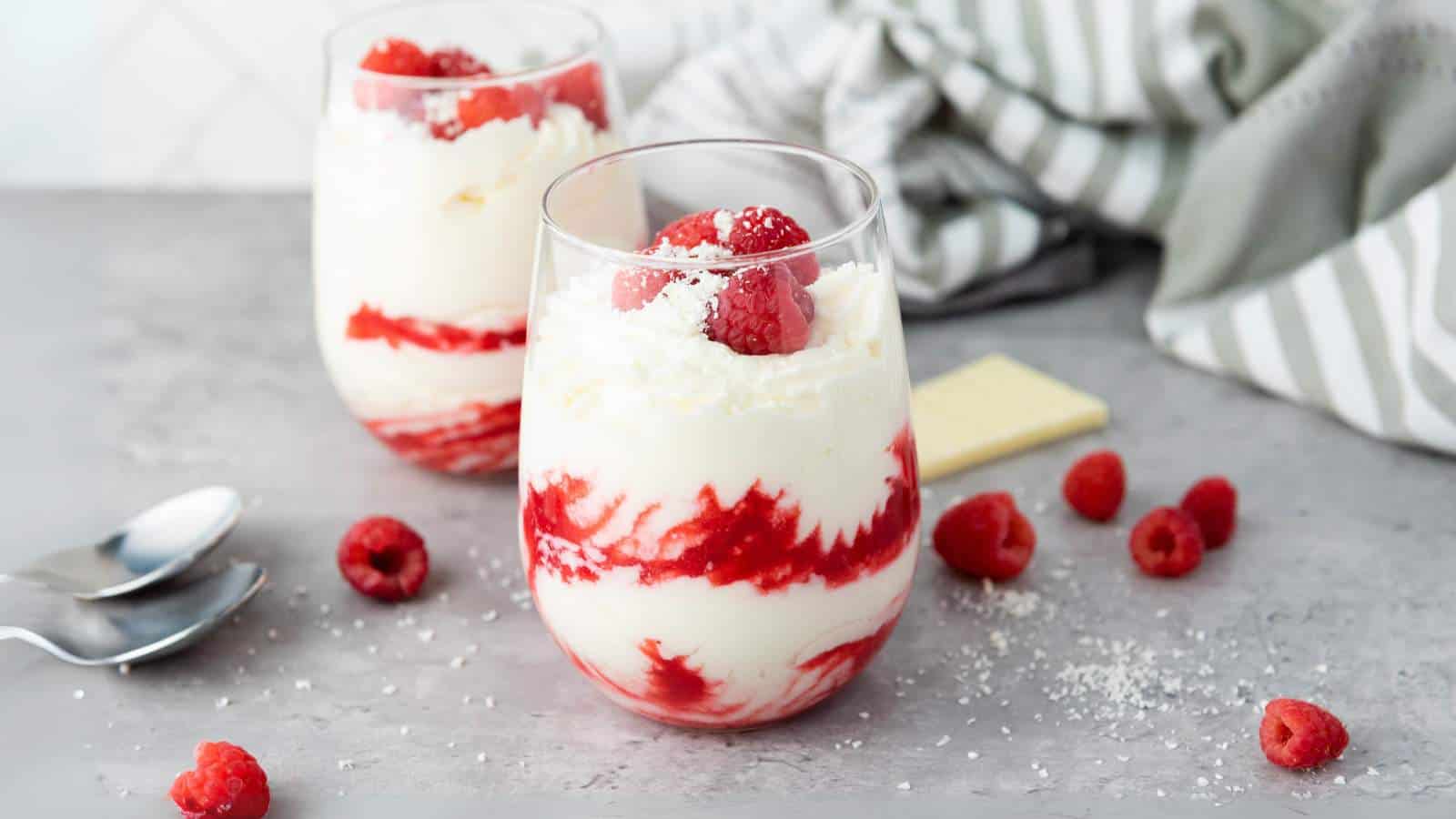 Two glasses of layered raspberry and cream dessert garnished with raspberries and white chocolate shavings, with extra raspberries and spoons on a gray surface.