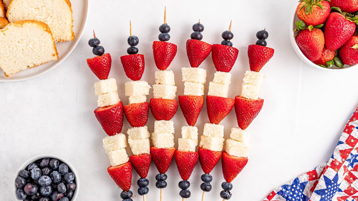 Fruit and cake skewers arranged on a white surface with strawberries, blueberries, and cubes of pound cake; bowls of fruit and cake slices are nearby.