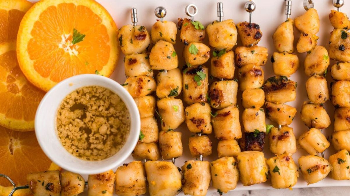 Grilled chicken skewers are arranged on a plate next to orange slices and a small bowl of crushed seasoning or dip.