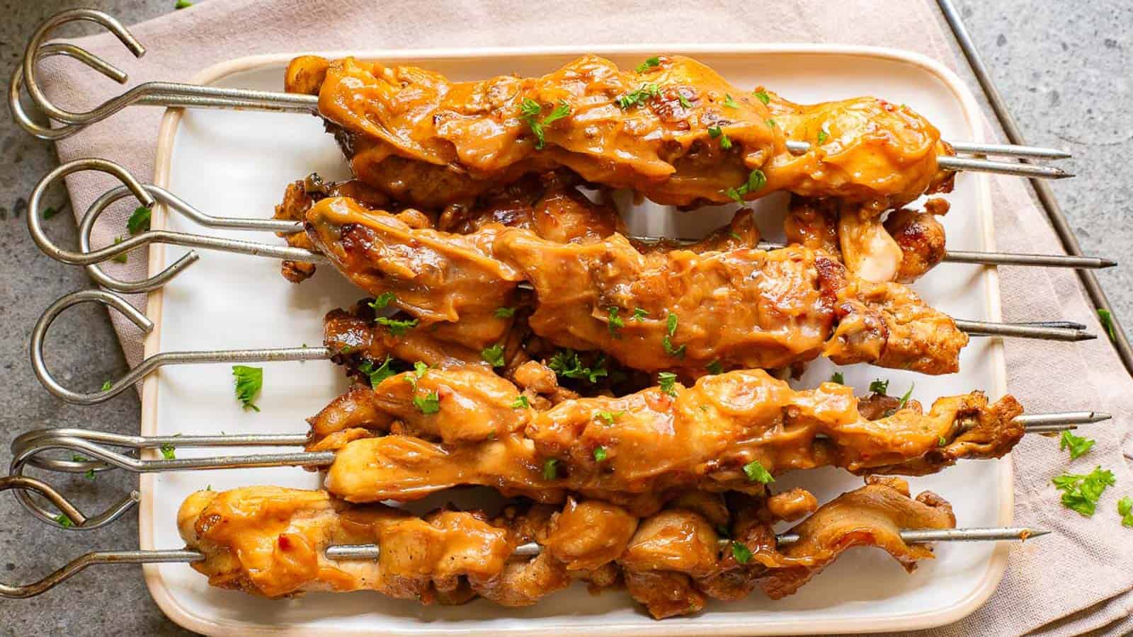 Five skewers of grilled chicken are arranged on a rectangular white plate, covered in a creamy orange sauce and garnished with chopped herbs.