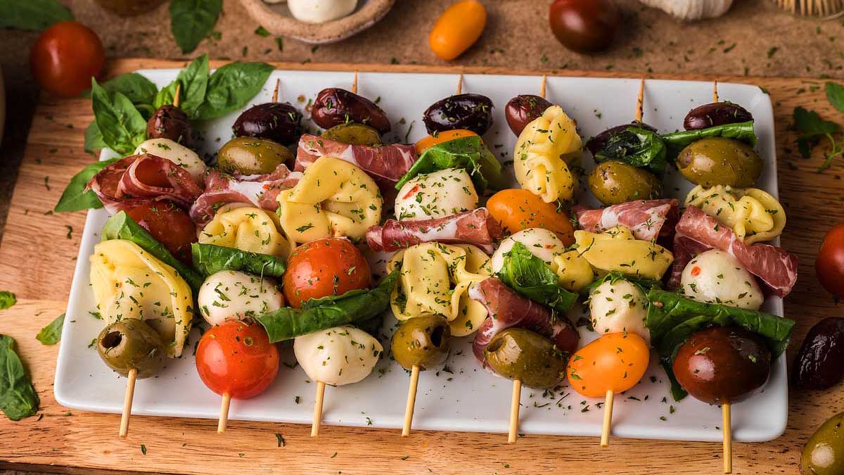 A white plate holds skewers with tortellini, cherry tomatoes, mozzarella balls, olives, basil leaves, and cured meats, garnished with herbs.