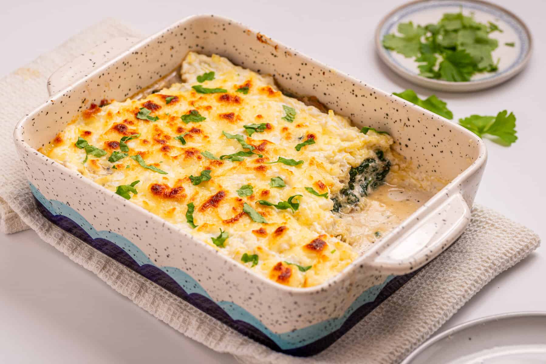 A baked casserole dish containing a golden-brown, cheesy lasagna, garnished with chopped parsley, sits on a white towel with a plate of fresh herbs nearby.