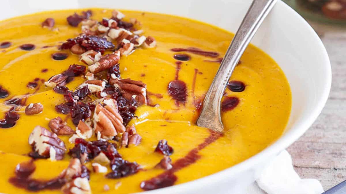 A bowl of creamy orange soup topped with chopped nuts, dried cranberries, and a drizzle of dark sauce, with a spoon resting inside.