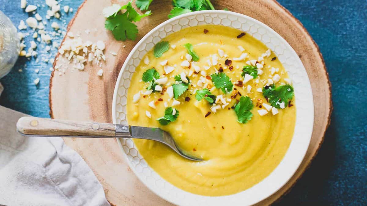 A bowl of creamy yellow soup garnished with chopped nuts, cilantro, and red pepper flakes, with a spoon resting inside, placed on a wooden board.