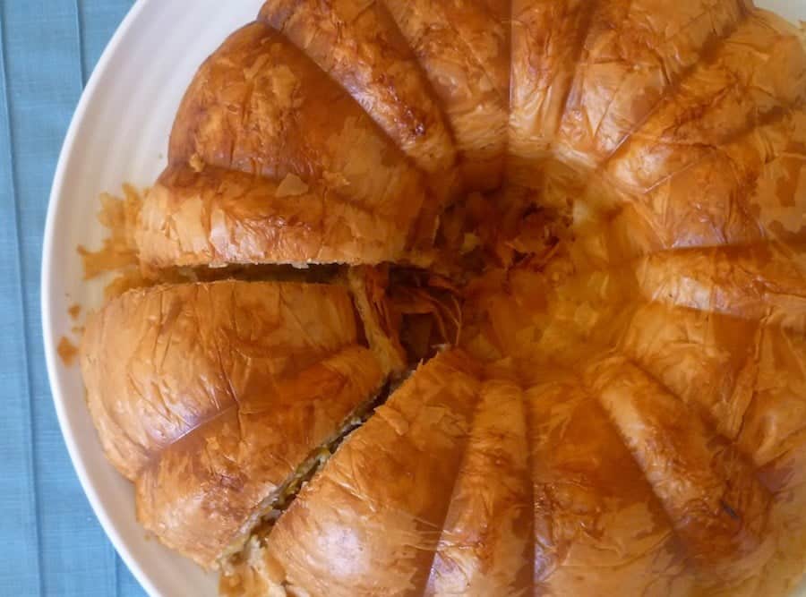 A round pie made of golden, flaky layered pastry is sliced and served on a white plate.