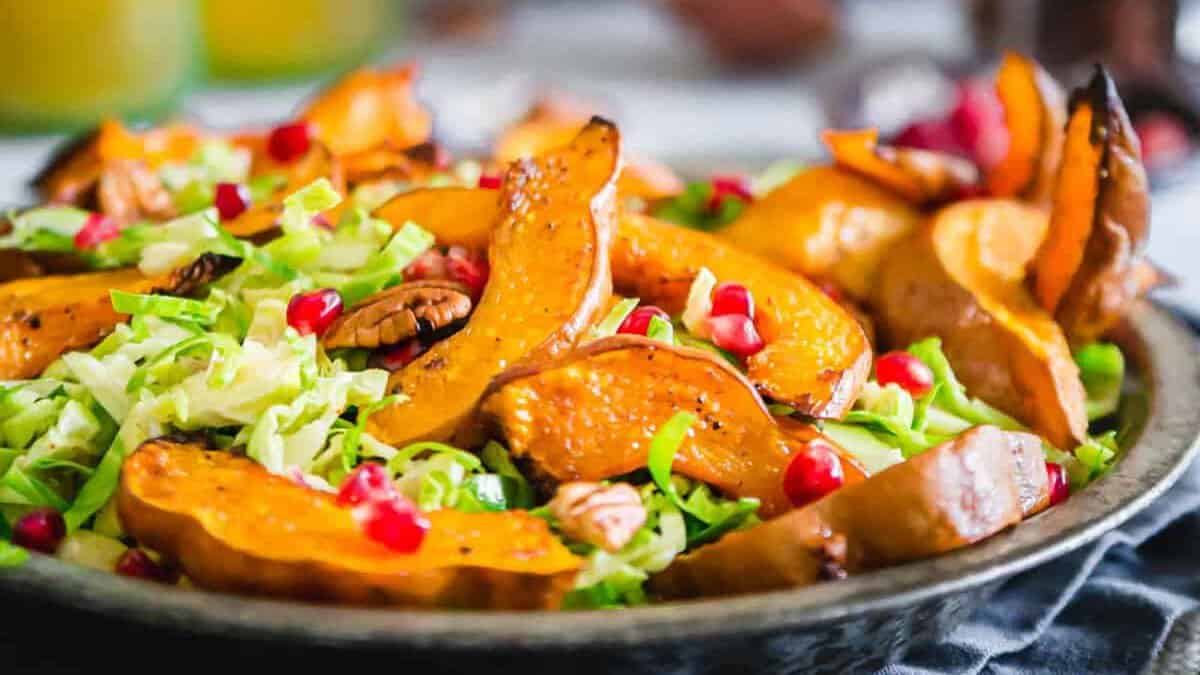A plate of roasted pumpkin slices, shredded lettuce, pecans, and pomegranate seeds on a dark dish.