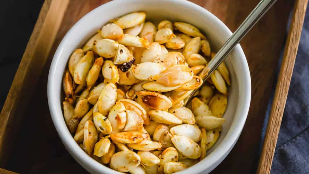 A white bowl filled with seasoned, roasted pumpkin seeds, with a metal spoon resting inside.