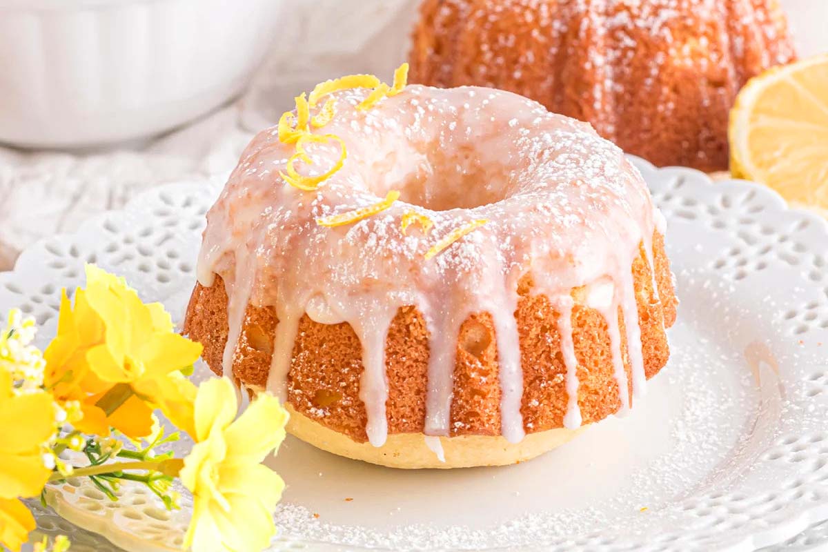 A mini bundt cake with white icing and lemon zest on top sits on a white plate, with yellow flowers and a lemon slice nearby.