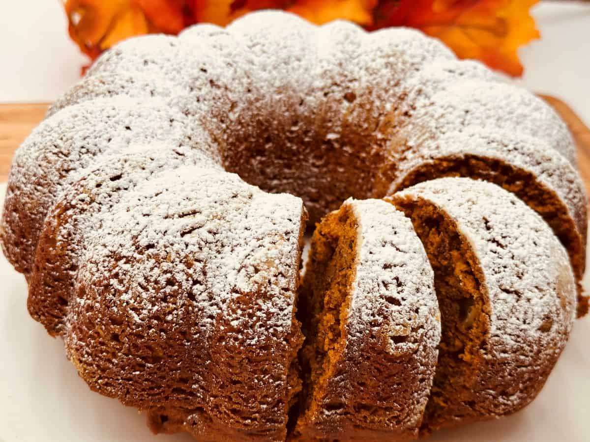 A bundt cake dusted with powdered sugar, with two slices cut, sits on a white surface. Autumn leaves are visible in the background.