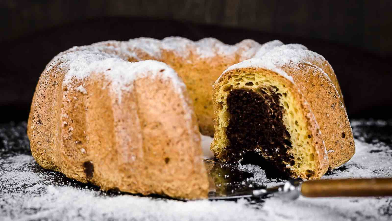 A sliced marble bundt cake with powdered sugar on top sits on a dark surface, with a serving knife partially under the cut piece.