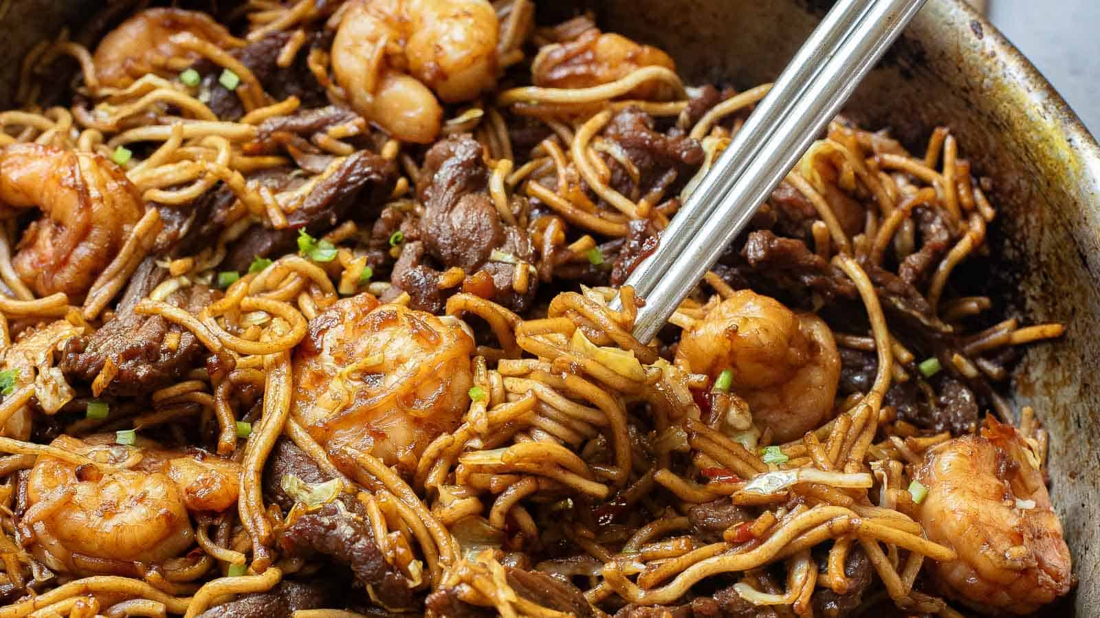 A close-up of stir-fried noodles with shrimp, beef, and chopped green onions, shown with a pair of metal chopsticks.
