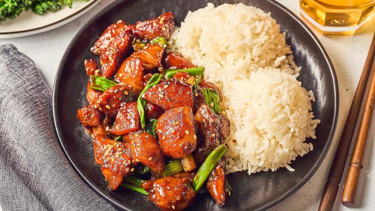 A black plate with glazed chicken pieces, green onions, and two servings of white rice. Chopsticks and a glass of drink are nearby.