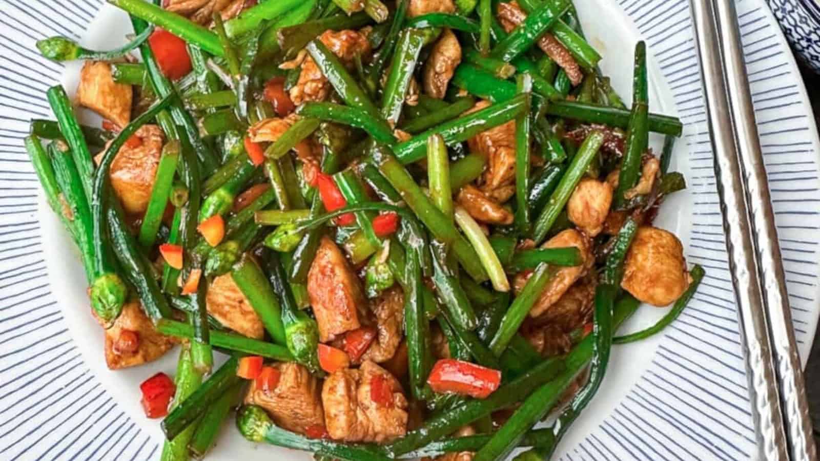 A plate of stir-fried chicken with green garlic shoots, red bell peppers, and a savory sauce, served on a white dish with metal chopsticks.
