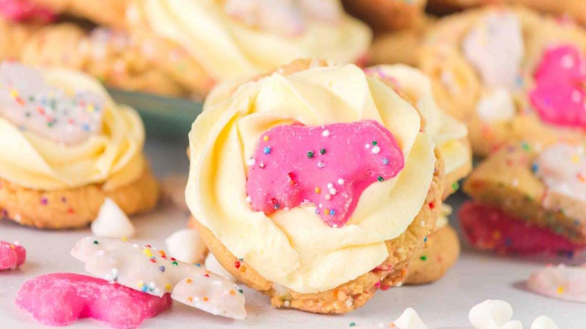 Frosted animal cookies with pink and white icing and sprinkles are placed on top of swirled vanilla frosting cookies, surrounded by more cookies and sprinkles.