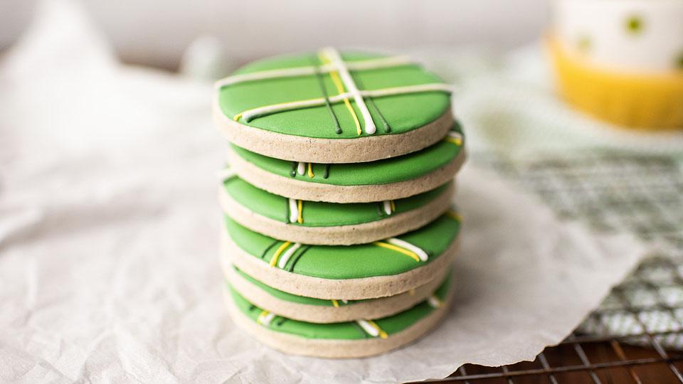 A stack of round sugar cookies with green icing and white, yellow, and black striped decorative lines sits on parchment paper.