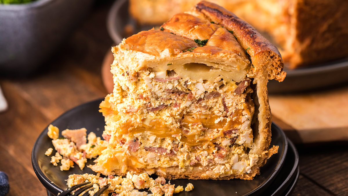 A close-up of a thick slice of savory meat and egg pie with golden-brown crust, served on a dark plate with a fork.