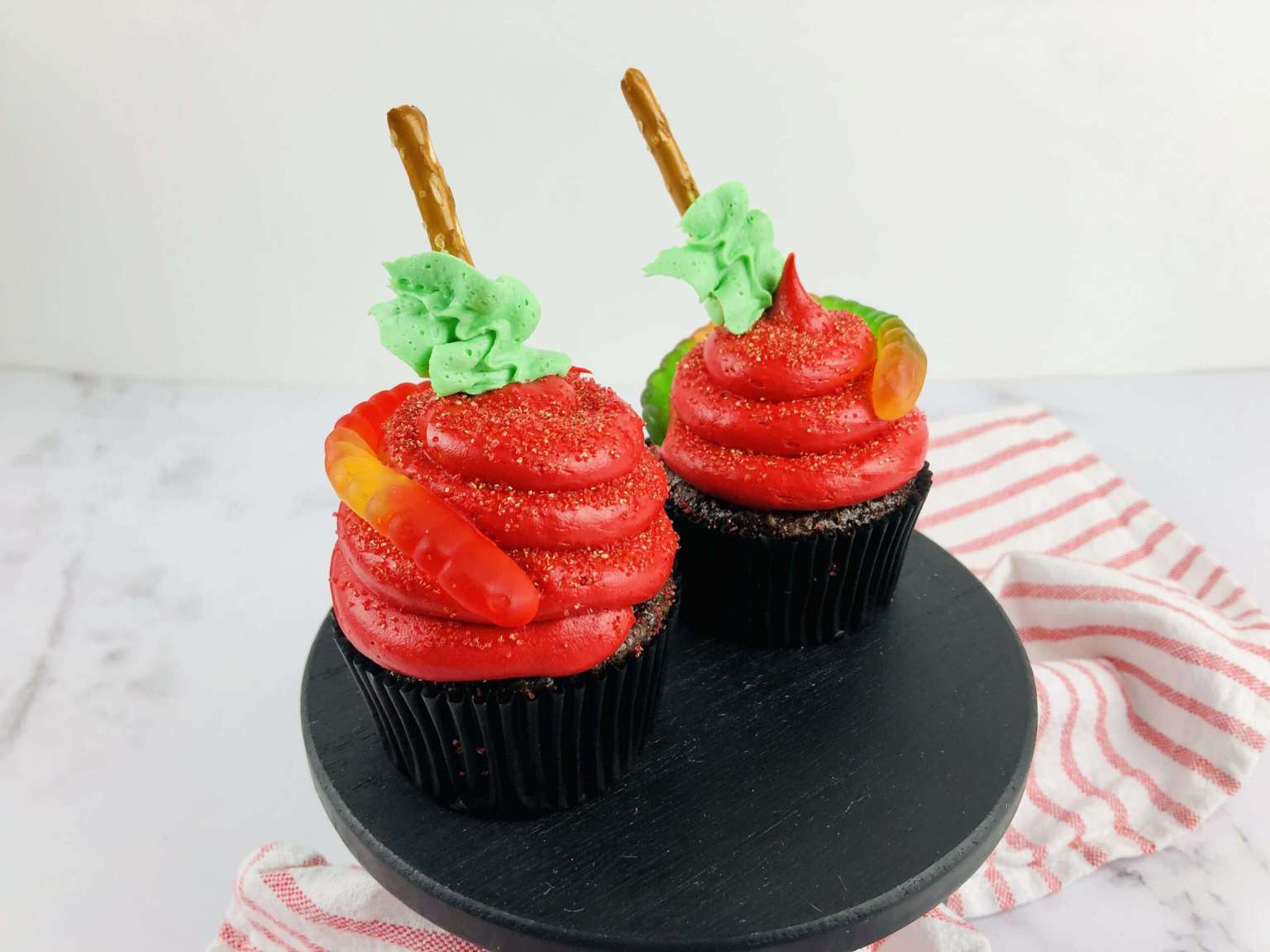 Two chocolate cupcakes with red frosting, green icing, gummy worms, and pretzel sticks on top, placed on a black stand with a striped cloth underneath.