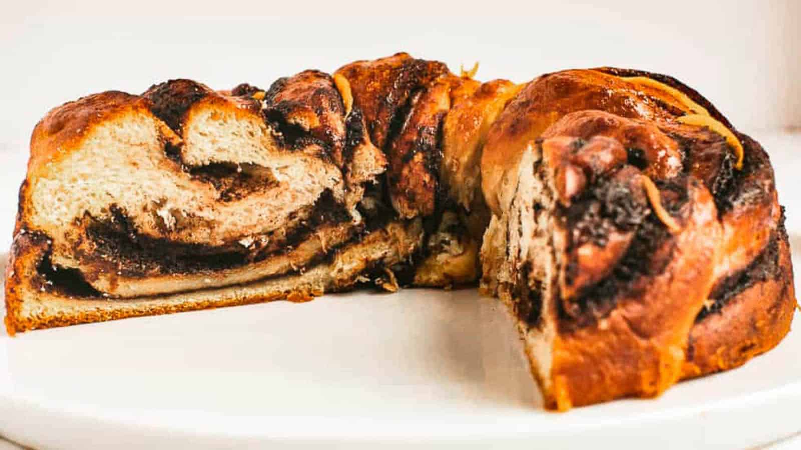 A round loaf of babka bread, with a slice removed to reveal chocolate swirls inside, sits on a white surface.