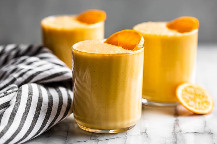 Three glasses of orange smoothie, each garnished with a persimmon slice, sit on a marble surface next to a striped cloth.
