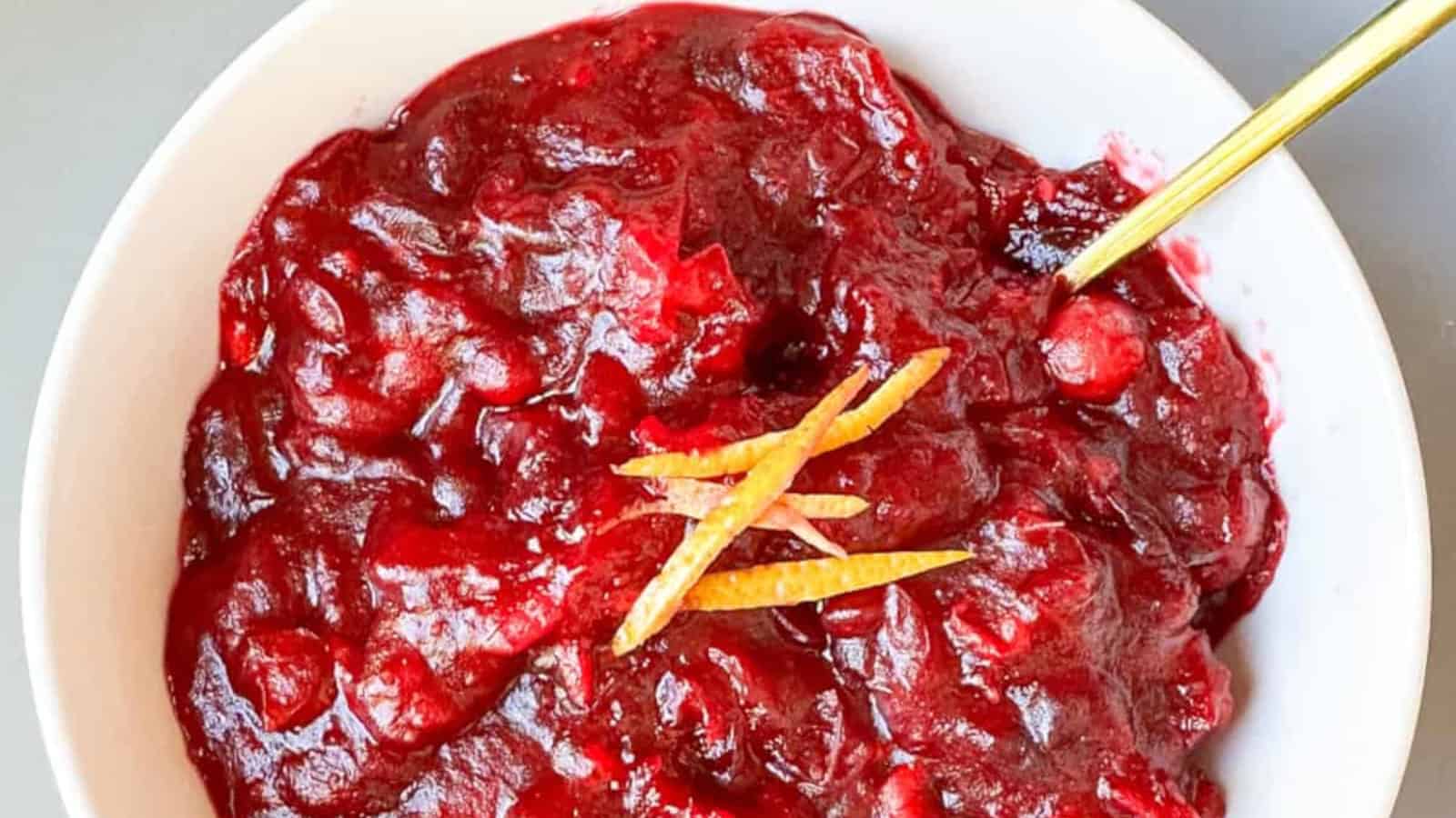 A bowl of cranberry sauce with chunks of fruit, topped with thin slices of orange peel, and a gold spoon resting inside.
