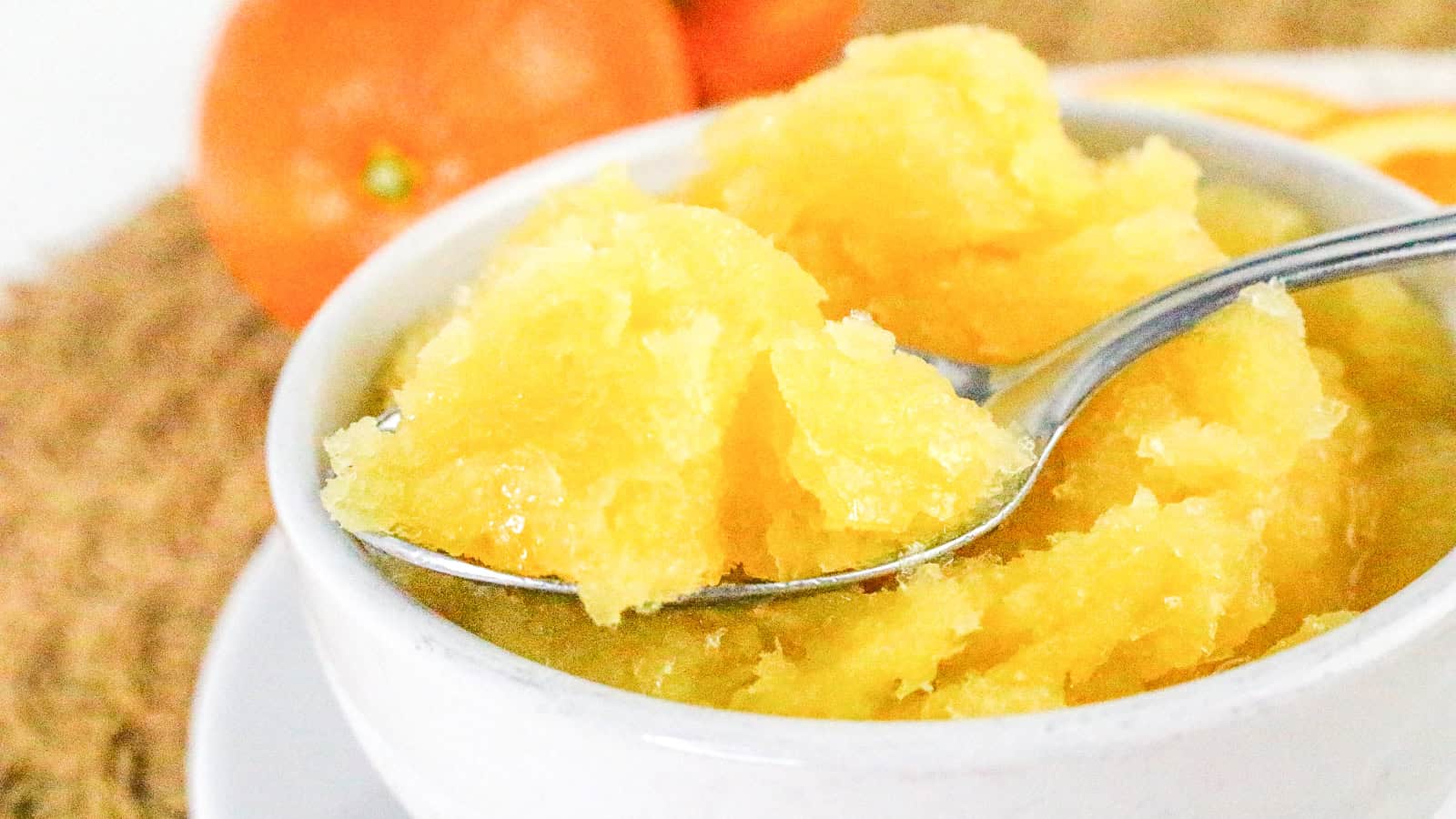 A close-up of a spoonful of orange sorbet being lifted from a bowl, with fresh oranges in the background.