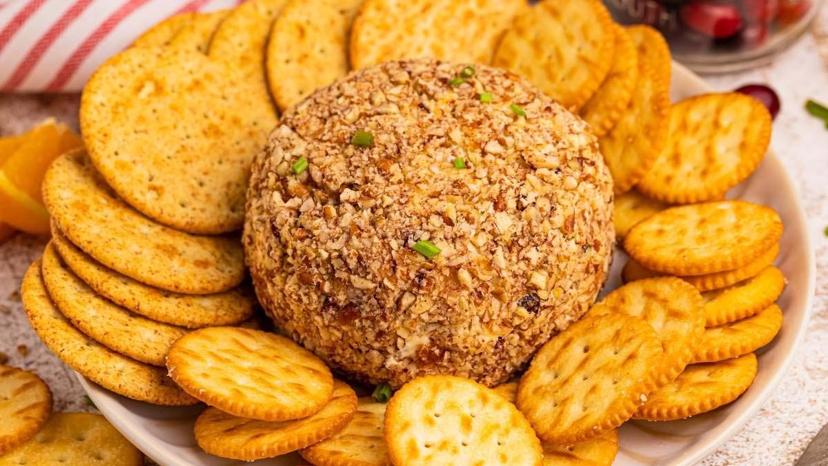 A round cheese ball coated in chopped nuts sits on a plate surrounded by assorted round crackers.