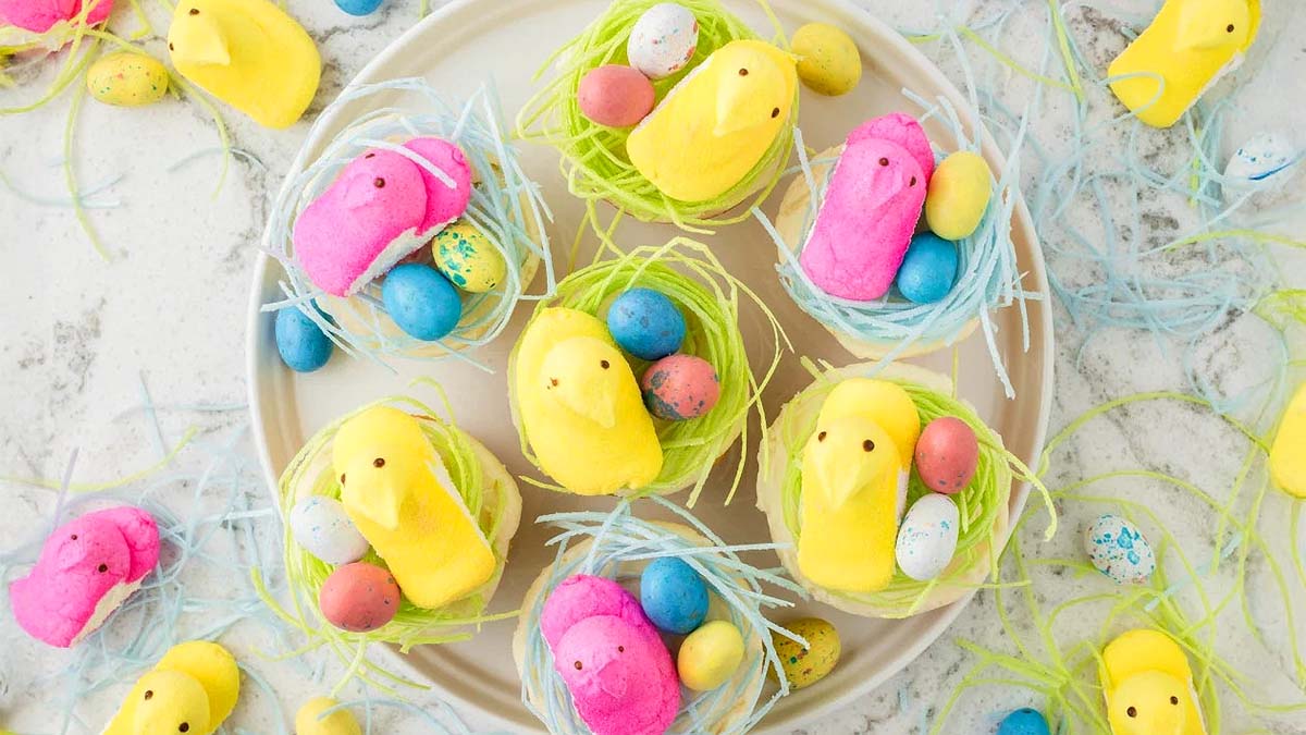 Colorful marshmallow chicks and candy eggs are arranged on a plate and scattered on a surface with blue and green paper grass, resembling Easter nests.