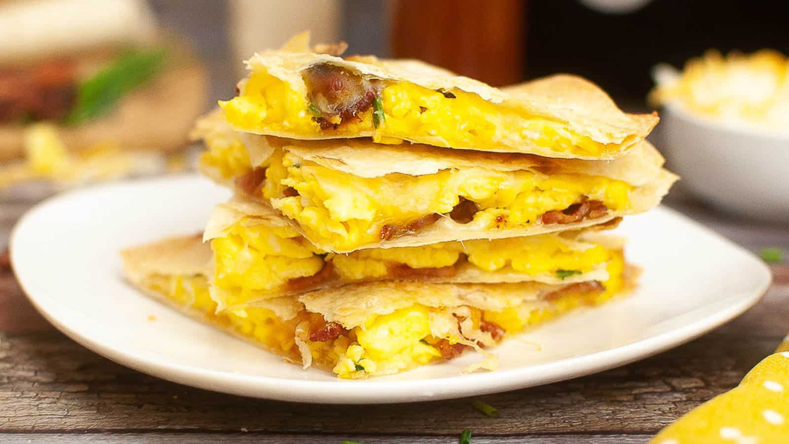 A stack of breakfast quesadillas filled with scrambled eggs and bacon is served on a white plate.