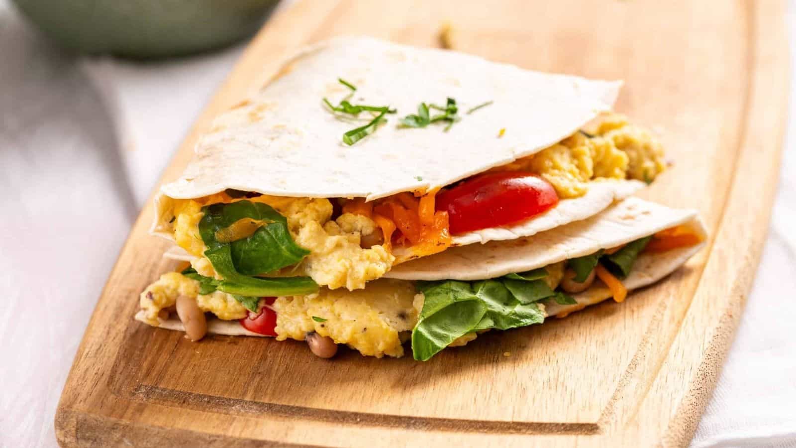 A folded tortilla filled with scrambled eggs, spinach, cherry tomatoes, and shredded cheese sits on a wooden cutting board.