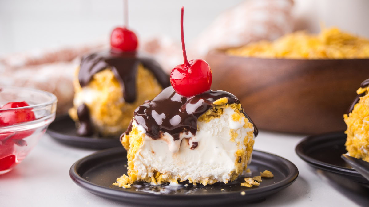 A dessert consisting of a scoop of fried ice cream topped with chocolate sauce and a maraschino cherry on a black plate.