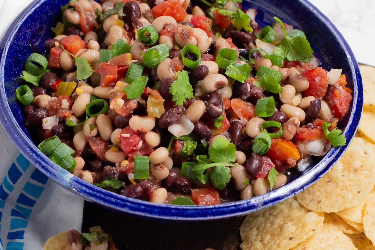 A blue bowl filled with bean salsa made of black beans, black-eyed peas, diced tomatoes, cilantro, and green onions, next to tortilla chips.