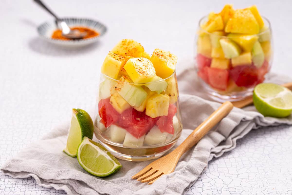 Two glasses filled with layered fruit pieces, including pineapple, watermelon, and melon, garnished with chili powder, with lime wedges and a wooden fork on a cloth napkin nearby.